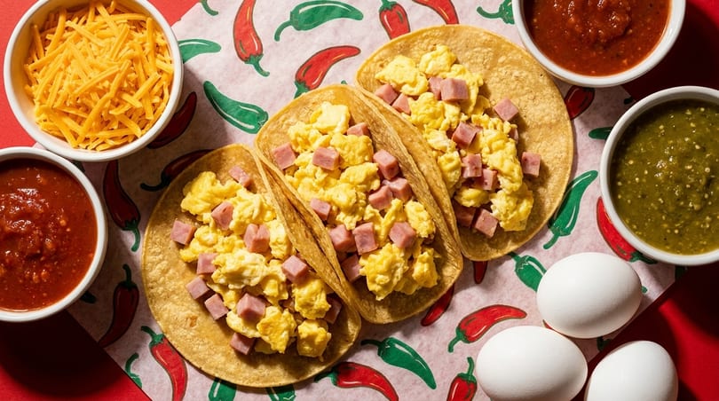 Three breakfast tacos with scrambled eggs and ham, alongside salsa, shredded cheese, and raw eggs.