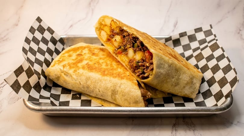 Two burritos, one cut open to show a filling of ground meat, potatoes, and vegetables.