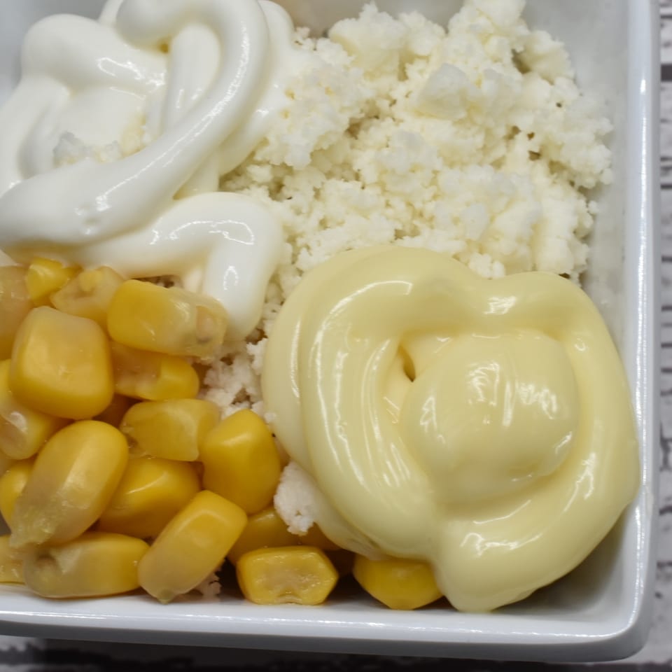 A white bowl filled with corn kernels, crumbled white cheese, and two swirls of mayonnaise.