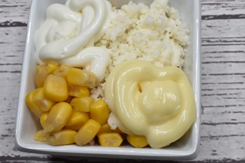 A white bowl filled with corn kernels, crumbled white cheese, and two swirls of mayonnaise.