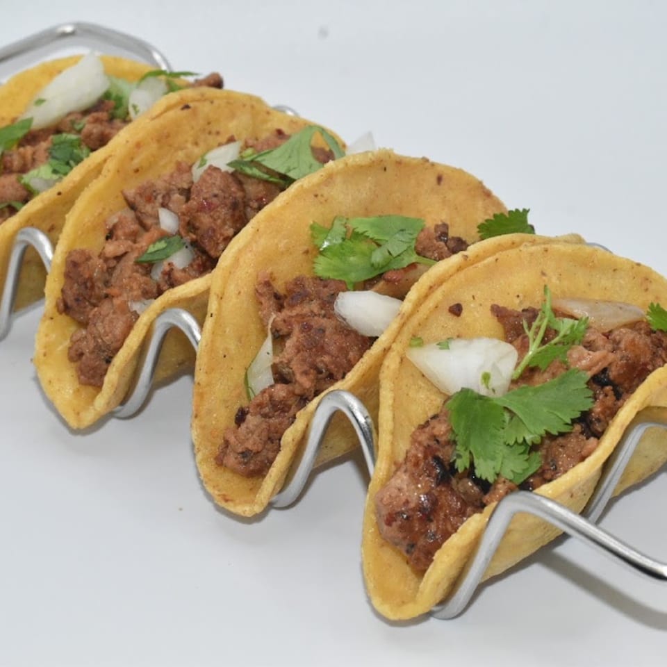 Four street tacos with meat, onions, and cilantro in a metal holder on a white surface.
