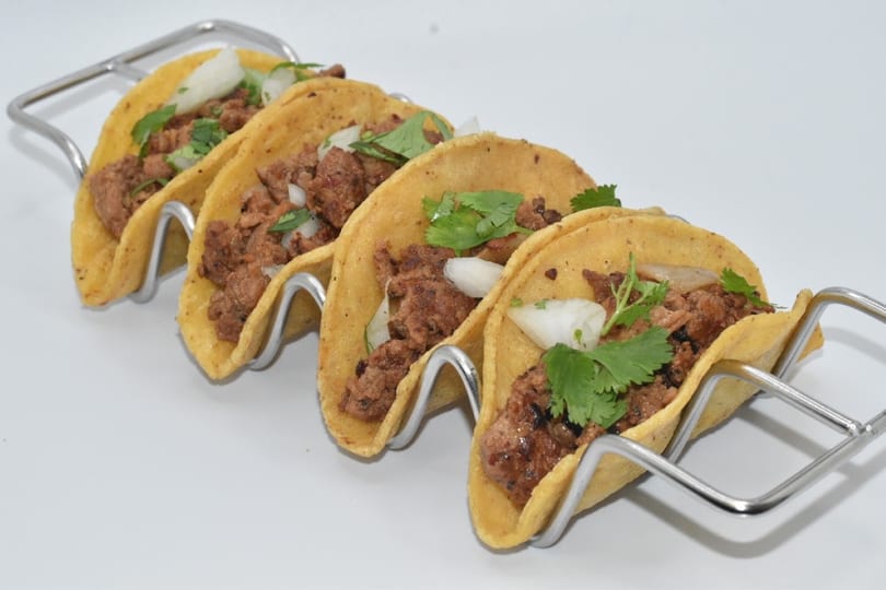 Four street tacos with meat, onions, and cilantro in a metal holder on a white background.