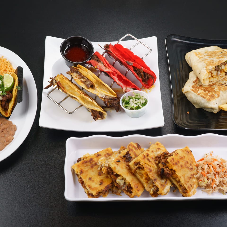 A black table displays various Mexican dishes: tacos, burritos, quesadillas, and stuffed peppers.