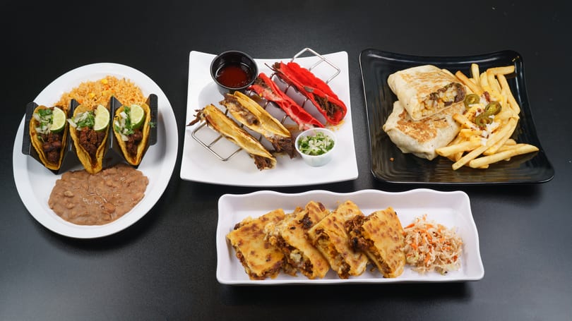 A black table displays various Mexican dishes: tacos, burritos, quesadillas, and stuffed peppers.